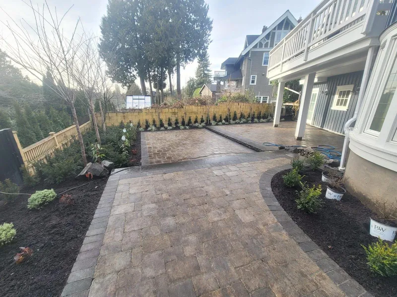 Paver patio and walkway with a dark border, new planting beds and a yellow brick retaining wall behind a house