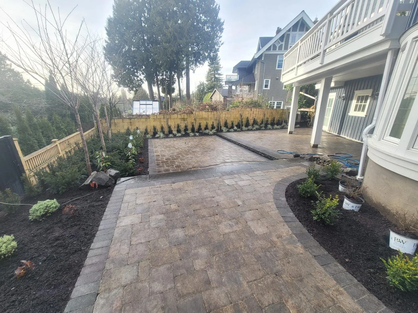 Paver patio and walkway with a dark border, new planting beds and a yellow brick retaining wall behind a house