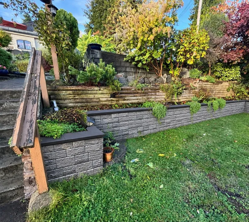 New split-face block retaining wall with a dark cap holding a planted slope above a lawn, beside timber steps
