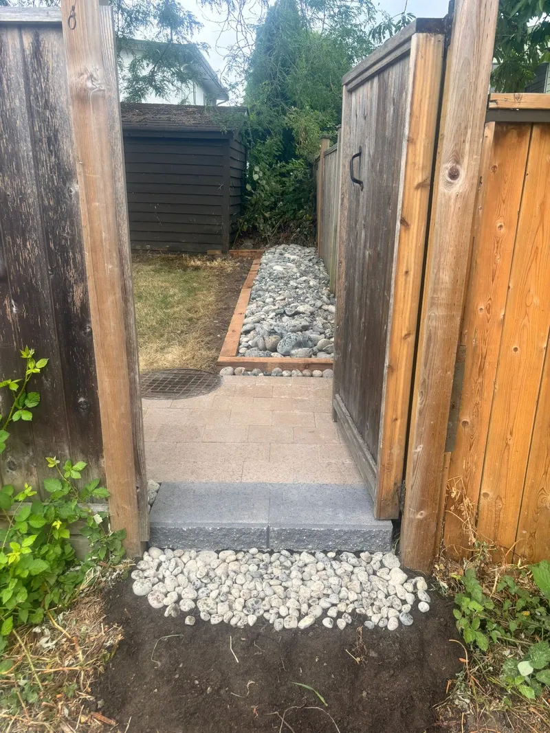 New paver path with river rock edging leading through a wood gate